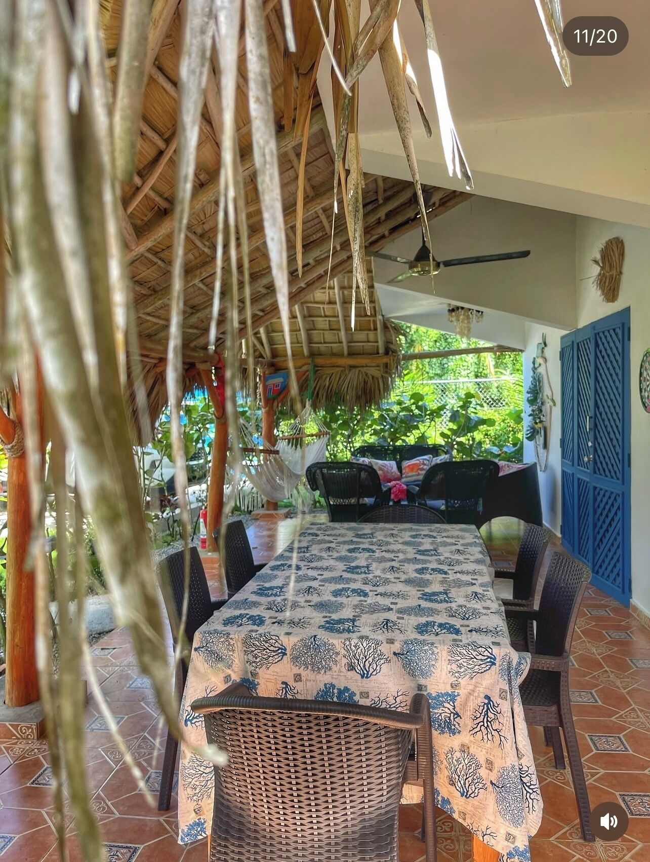 A covered outdoor dining area with a table and chairs, a thatched roof, and a view of greenery.