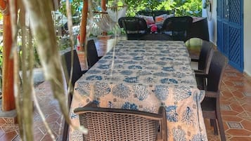 A covered outdoor dining area with a table and chairs, a thatched roof, and a view of greenery.