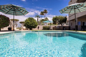 A clear swimming pool with two striped umbrellas, palm trees, and a building in the background. - Spacious 5-bedroom house in serene Lake Havasu with pool, and Lake view (Lake Havasu City)