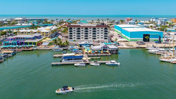 A marina with boats docked, a large building with a blue facade, and a waterfront restaurant area. - Beachfront Escape with Pool & Island Fun Steps from the Sand - Sand Caper 509 - Roelens (Fort Myers Beach)