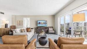A living room with a sofa, chairs, a coffee table, a TV, and a balcony with a table and chairs. - Beachfront Escape with Pool & Island Fun Steps from the Sand - Sand Caper 509 - Roelens (Fort Myers Beach)