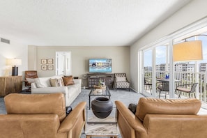 A living room with a sofa, chairs, a coffee table, a TV, and a balcony with a table and chairs. - Beachfront Escape with Pool & Island Fun Steps from the Sand - Sand Caper 509 - Roelens (Fort Myers Beach)