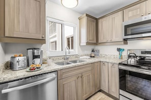 A kitchen with wooden cabinets, a stainless steel dishwasher, a microwave, and a stove. - Beachfront Escape with Pool & Island Fun Steps from the Sand - Sand Caper 509 - Roelens (Fort Myers Beach)