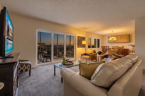A living room with a sofa, a coffee table, and a television. There is a balcony with a view of the city. - Beachfront Escape with Pool & Island Fun Steps from the Sand - Sand Caper 509 - Roelens (Fort Myers Beach)