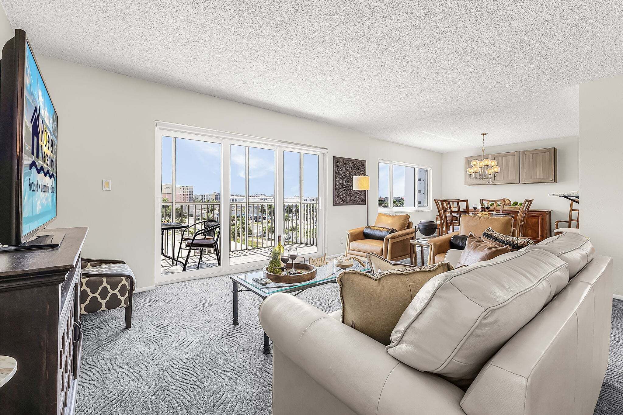 A spacious living room with a large sofa, a dining area with chairs, a television, and a balcony with a view.