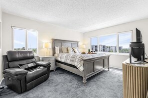 Une chambre à coucher avec un lit, un fauteuil en cuir, une table de chevet avec des lampes et une vue sur les immeubles à travers de grandes fenêtres.