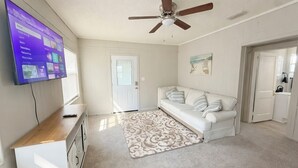 Una habitación con ventilador de techo, un sofá blanco, un televisor montado en la pared y una puerta que da a otra habitación.