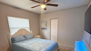 Un dormitorio con cama, mesita de noche, cómoda, ventilador de techo, ventana con persianas y puerta.