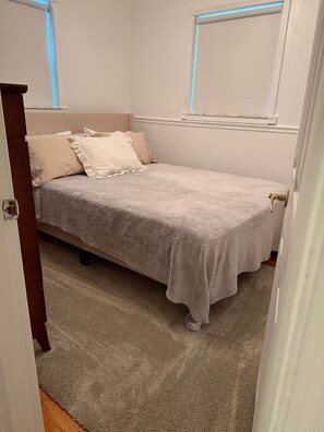 A bedroom with a single bed, a window with blinds, a carpeted floor, and a door.