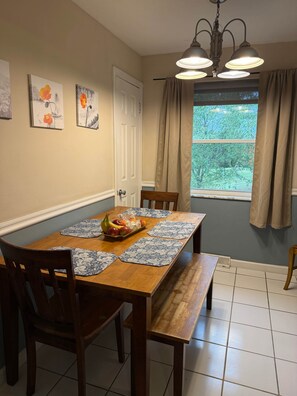 A dining area with a wooden table, blue placemats, and chairs. There are paintings on the wall, a window with curtains, and a chandelier.