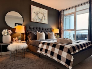 Une chambre à coucher avec un grand lit, deux lampes de chevet, un miroir rond, un tableau et une vue sur l’extérieur.