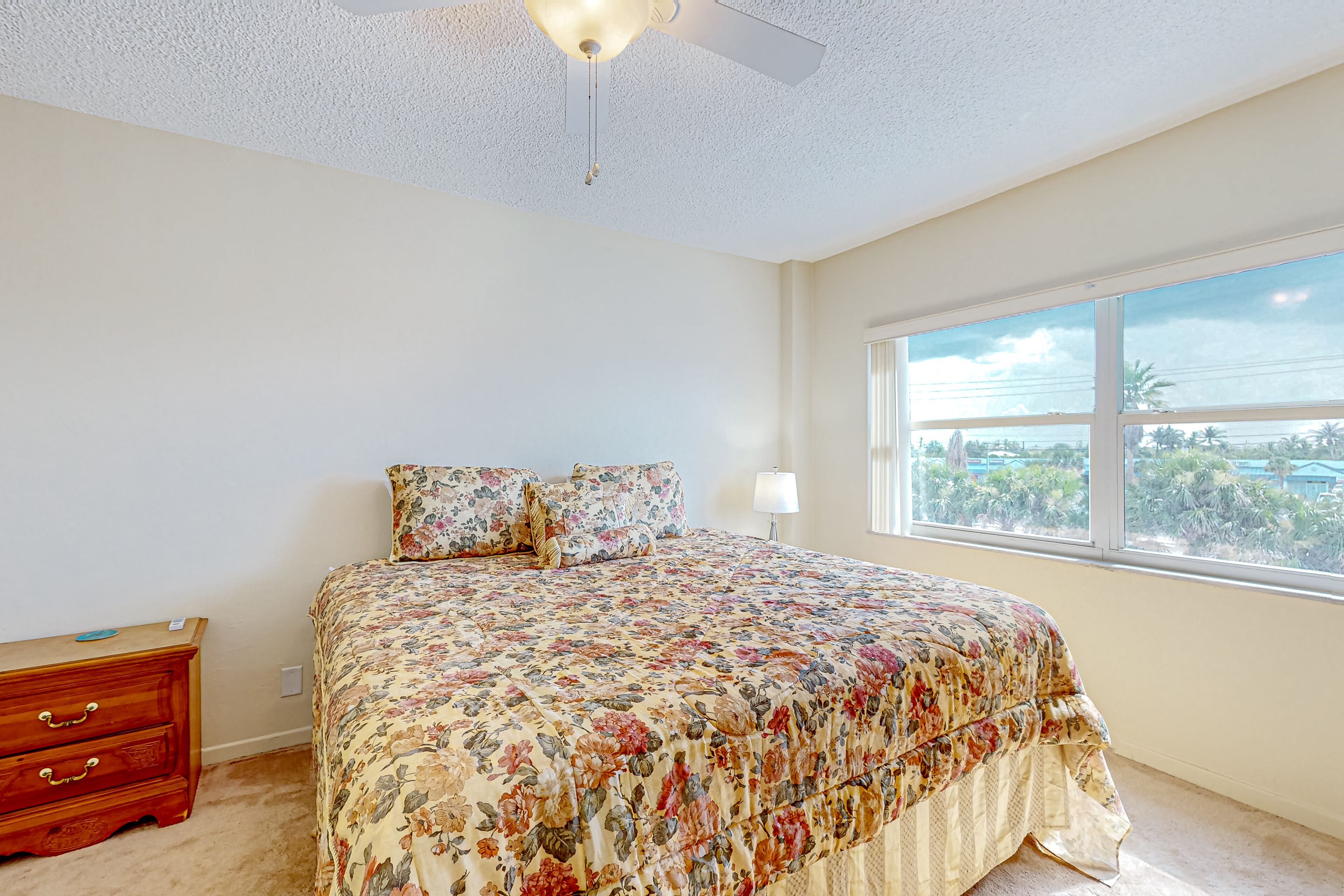 Quarto com colcha florida, mesa de cabeceira de madeira, ventilador de teto e janela com vista para palmeiras.