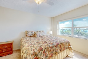 Quarto com colcha florida, mesa de cabeceira de madeira, ventilador de teto e janela com vista para palmeiras.
