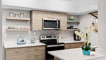 Uma cozinha moderna com micro-ondas, fogão e torradeira. Há armários de madeira, bancada branca e um vaso com flores.
