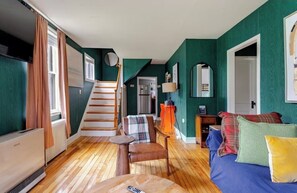 A room with a staircase, a bed with colorful pillows, a small table with a remote control, and a radiator. - Historic Schoolhouse on river in enchanting Keene Valley Adirondacks (Keene Valley)