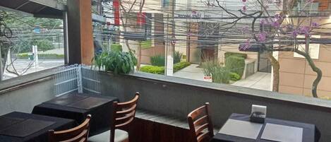 Una sala da pranzo interna con tavoli e sedie, ampie finestre che offrono vista su edifici e alberi.