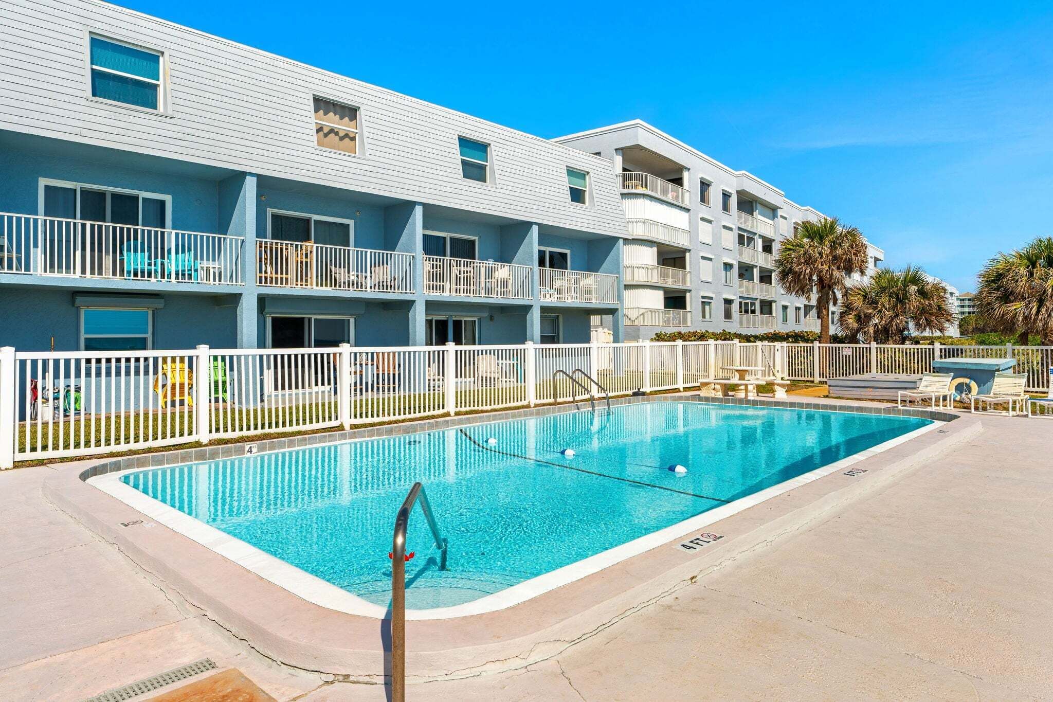 A swimming pool with a white fence, a multi-story building with balconies, and palm trees.