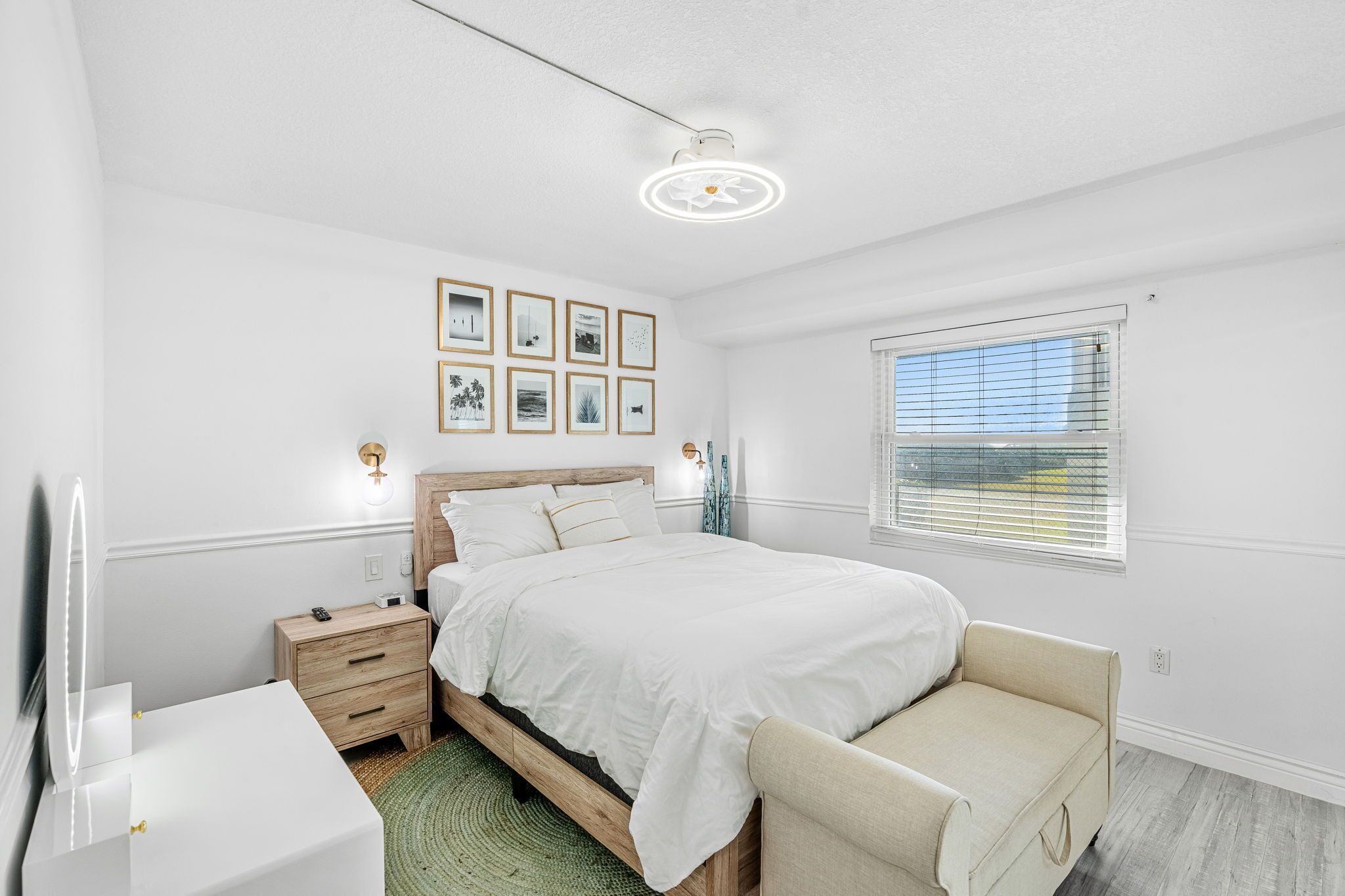 A bedroom with a bed, bedside tables, a bedside lamp, a window with blinds, and framed pictures on the wall.