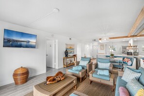A modern living room with wicker furniture, a large painting, and a kitchen area with a hanging light fixture. - Seabreakers 2 (Satellite Beach)