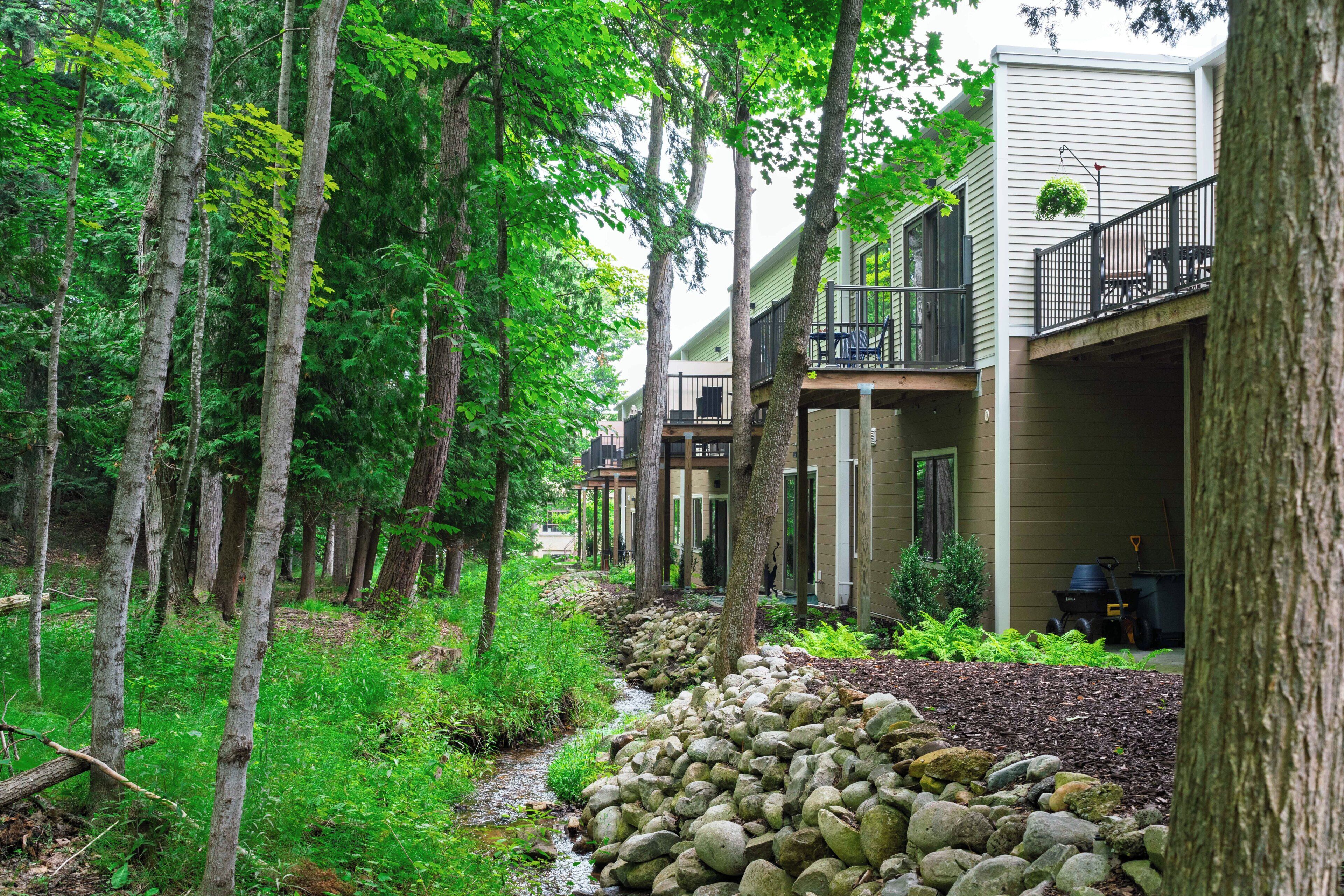 A residential area with a pathway lined with trees and a small stream. There are houses with balconies and a visible outdoor grill.