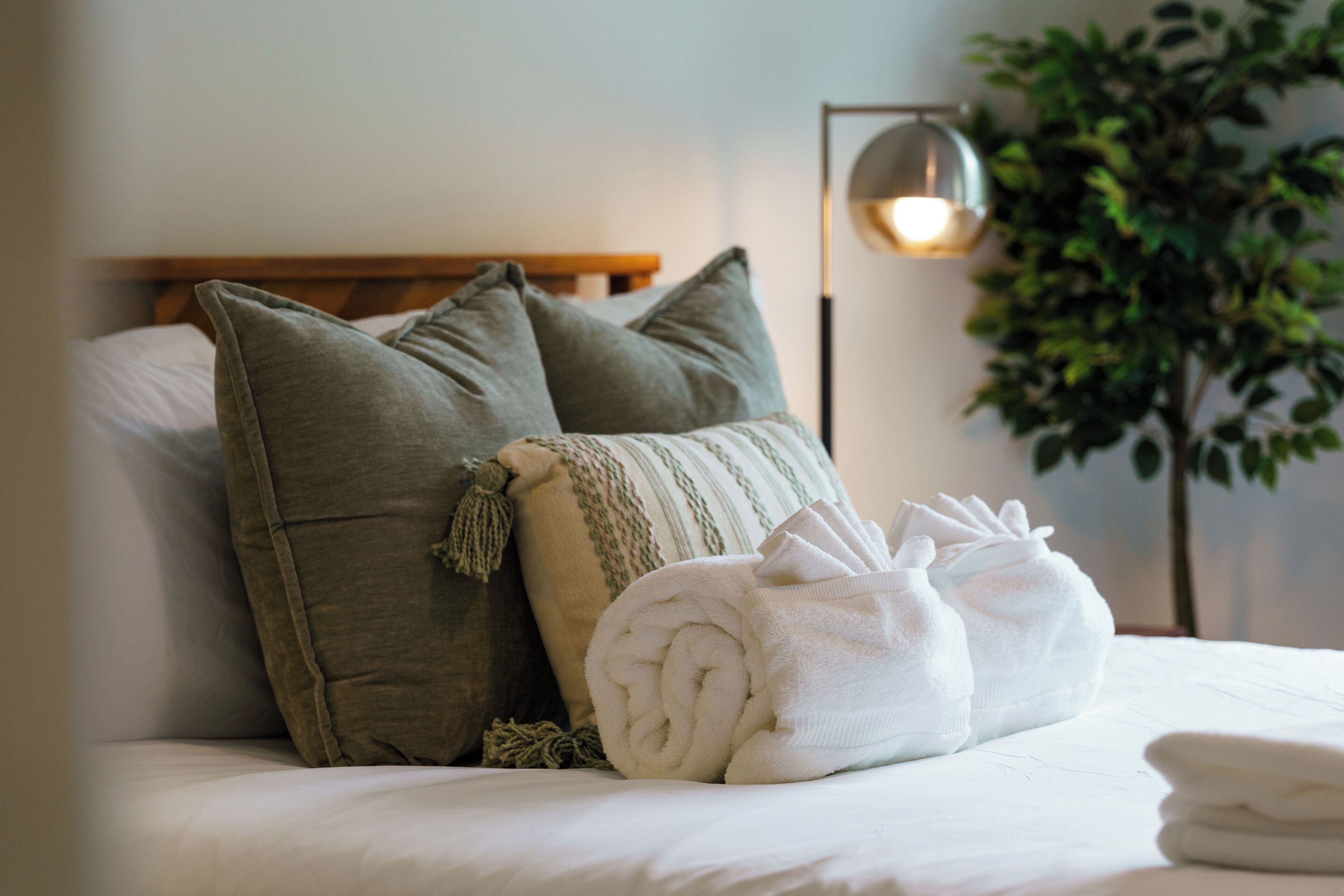 Una cama bien hecha con almohadas y una toalla enrollada, una lámpara de noche y una planta en maceta.