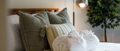 A neatly made bed with pillows and a rolled towel, a bedside lamp, and a potted plant.