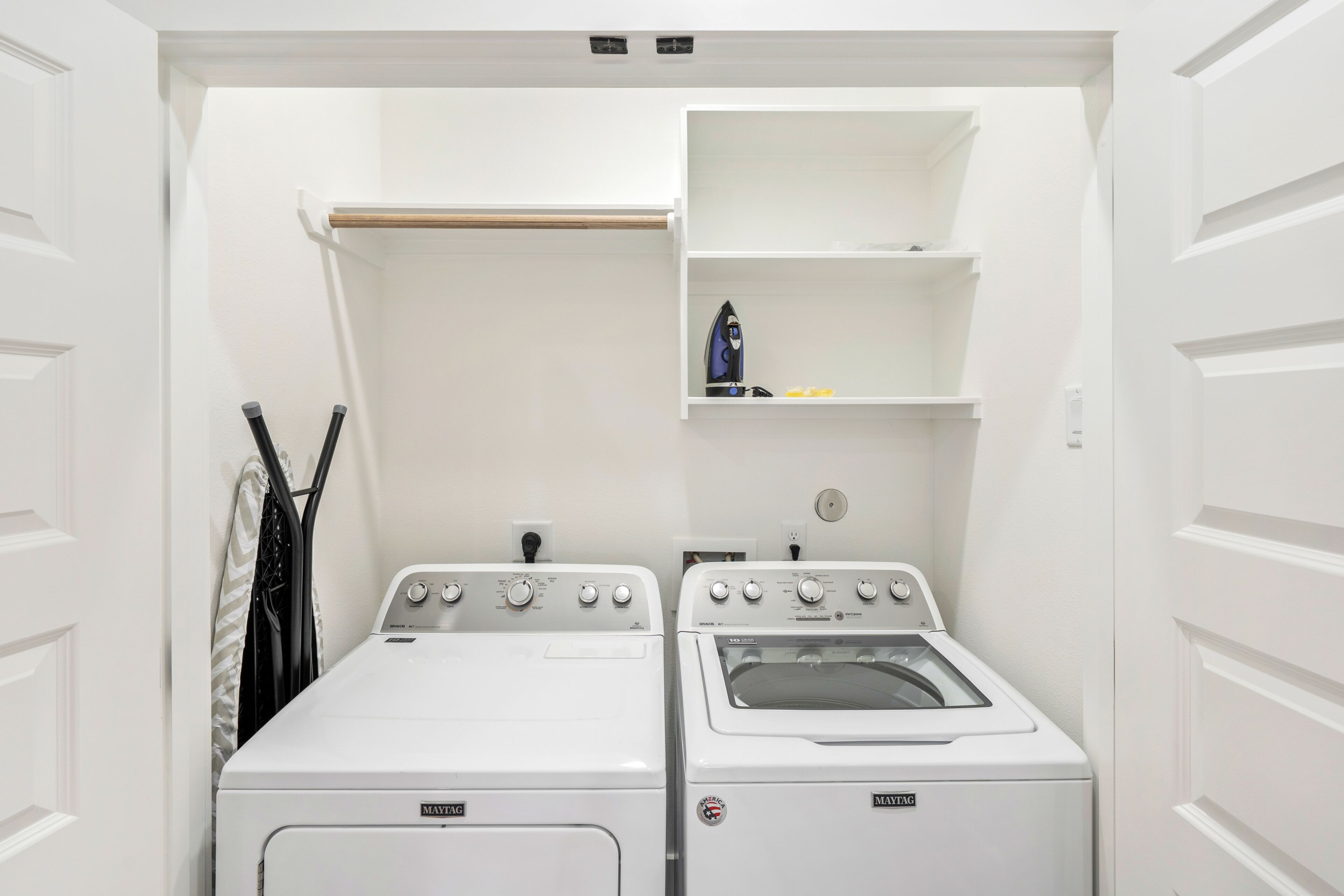 A compact laundry room with two Maytag washers and dryers, a drying rack, and a shelf with a small statue and a bottle.