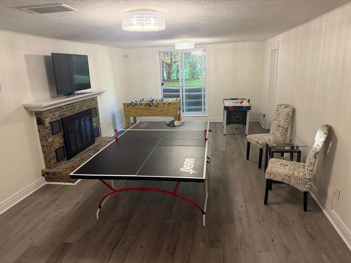 A room with a ping pong table, a fireplace, a television, and a window with a view of greenery.