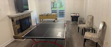A room with a ping pong table, a fireplace, a television, and a window with a view of greenery.