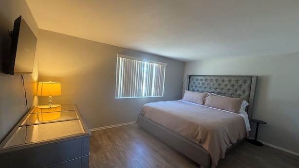A bedroom with a bed, a bedside table with a lamp, a window with blinds, and a wall-mounted TV. - New Port Richey Retreat- Sleeps 4 (New Port Richey)