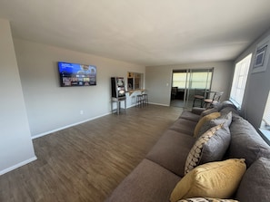 Amplia sala de estar con una televisión de pantalla plana grande montada en la pared, zona de cocina con barra y puerta corrediza de vidrio que da a un balcón.