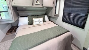 Una camera da letto con un letto, cuscini, una testiera, una finestra con persiane e una mensola a parete.