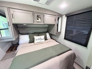 Ein Schlafzimmer mit Bett, Kissen, Kopfteil, einem Fenster mit Jalousien und einem an der Wand befestigten Regal.
