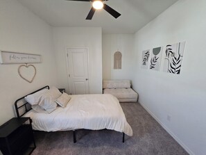 Ein Schlafzimmer mit Bett, Nachttisch, Deckenventilator, Tür und Wanddekorationen, darunter ein herzförmiger Bilderrahmen und Kunstwerk mit Blätermotiv.