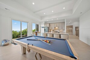 A pool table in a modern room with a kitchen and dining area, large windows, and a view of greenery. - High View: Brand-New 5,100 Sq Ft Nordic-Inspired Mountain Estate with Four-State Views (Banner Elk)