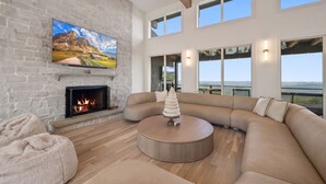 A modern living room with a fireplace, a large sectional sofa, a round coffee table, and a television mounted on a stone wall. - High View: Brand-New 5,100 Sq Ft Nordic-Inspired Mountain Estate with Four-State Views (Banner Elk)