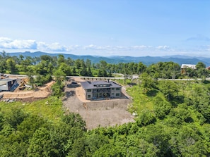 A large house with a spacious yard, surrounded by dense greenery and mountains in the background. - High View: Brand-New 5,100 Sq Ft Nordic-Inspired Mountain Estate with Four-State Views (Banner Elk)