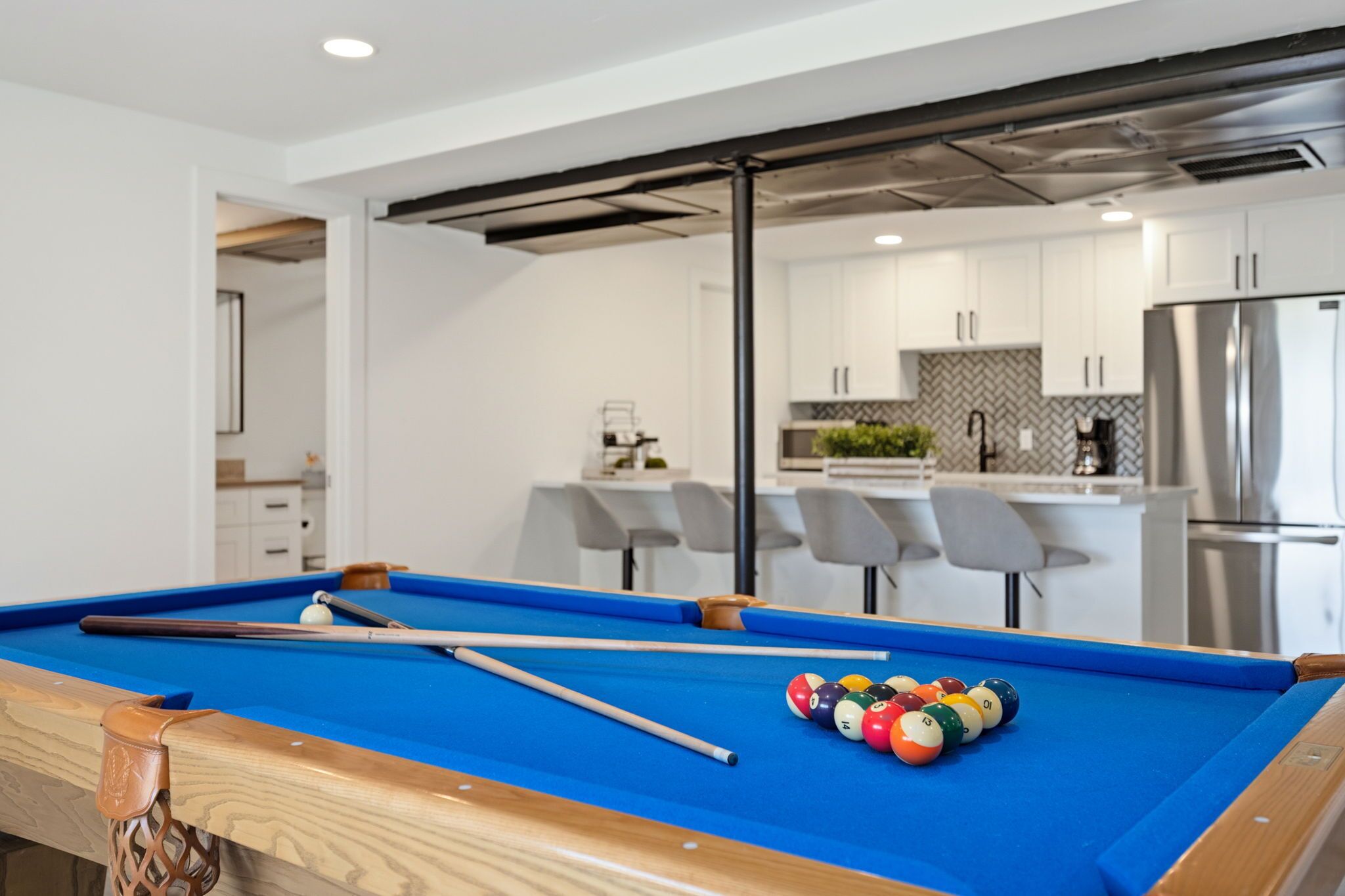 A pool table with pool cues and balls, a kitchen with white cabinets and stainless steel appliances, and a bar area with bar stools.