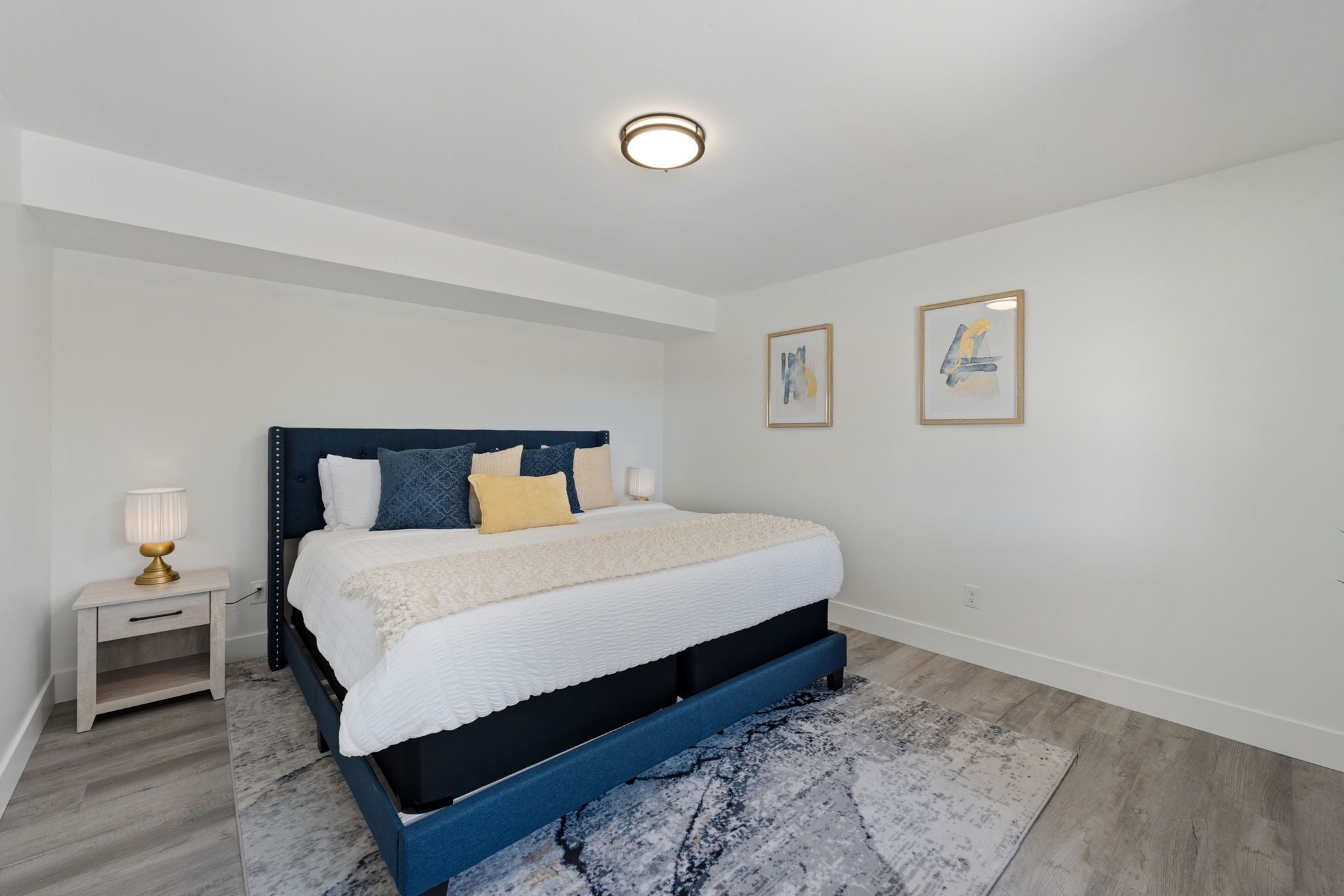 A bedroom with a bed, bedside table, lamp, and two framed artworks on the wall.