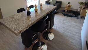 A modern kitchen with a granite countertop, bar stools, a wall clock, and a large window.