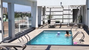 A rooftop swimming pool with a view of the city skyline, surrounded by lounge chairs and a potted plant.