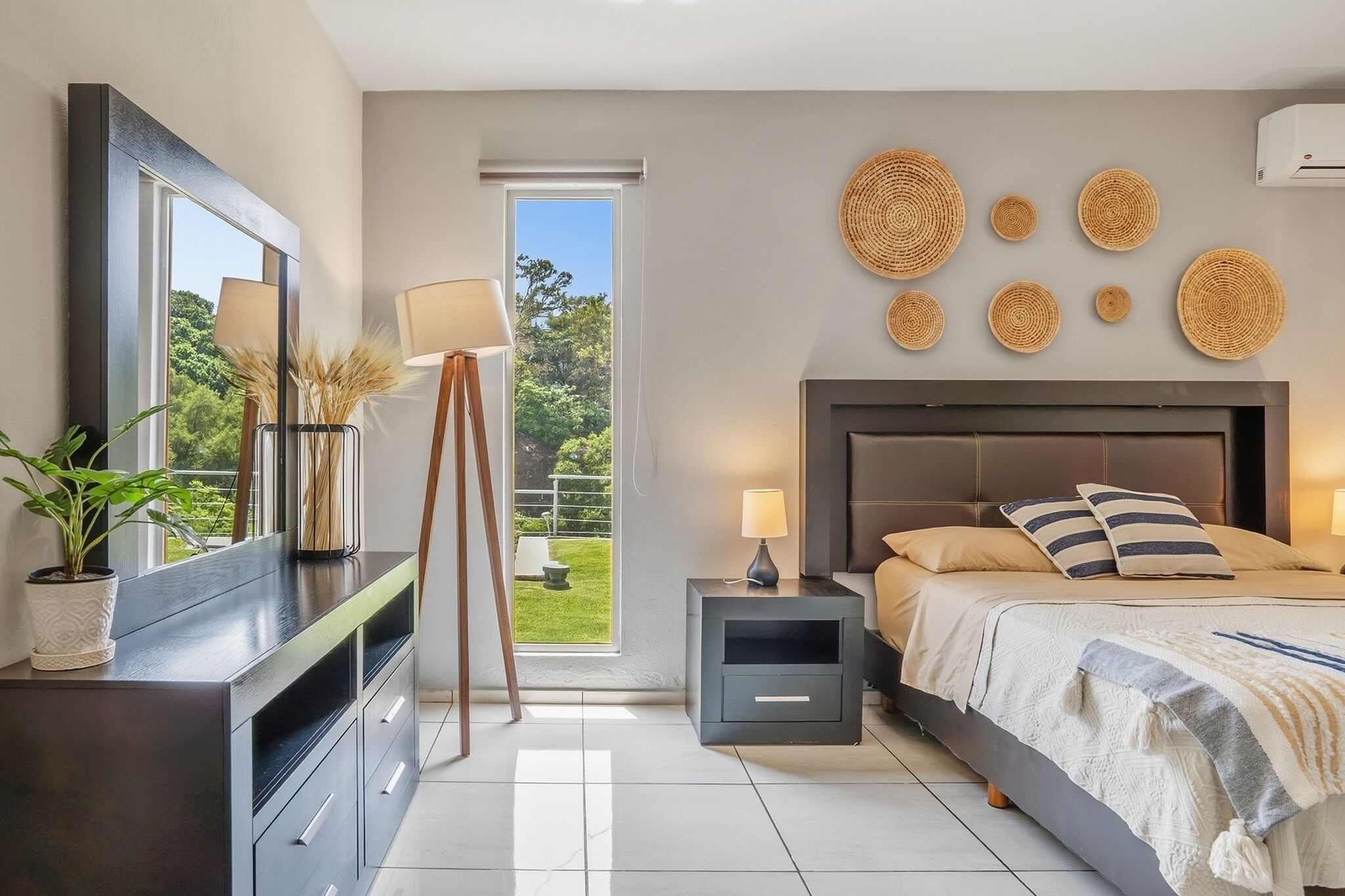 Un dormitorio moderno con cama, mesitas de noche, televisor y vistas al exterior con vegetación.