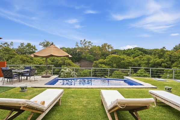 Zona de piscina con sillones de descanso, una mesa y una sombrilla.