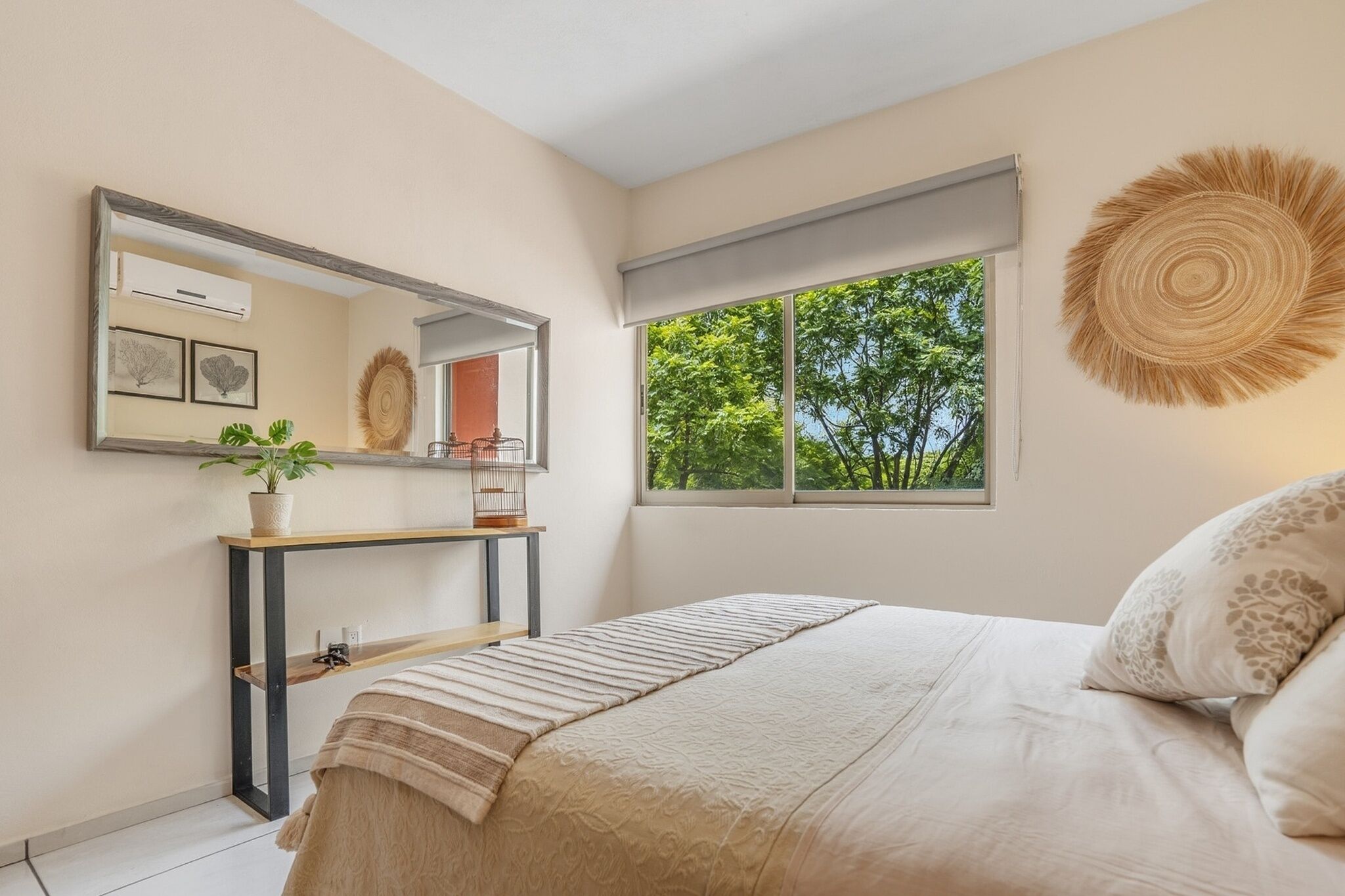 Une chambre à coucher avec un lit, une table de chevet ornée d’une plante, un miroir et une fenêtre donnant sur des arbres.