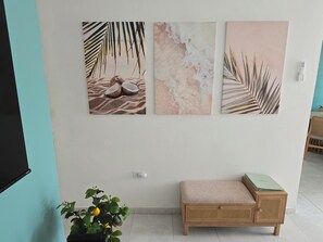 A wall with three framed artworks depicting beach scenes, a wooden bench with a cushion, and a potted plant with yellow flowers. - Cozy 3BR house with AC•Parking•Patio•Centric Location
Casa Jeyri (Cataño)