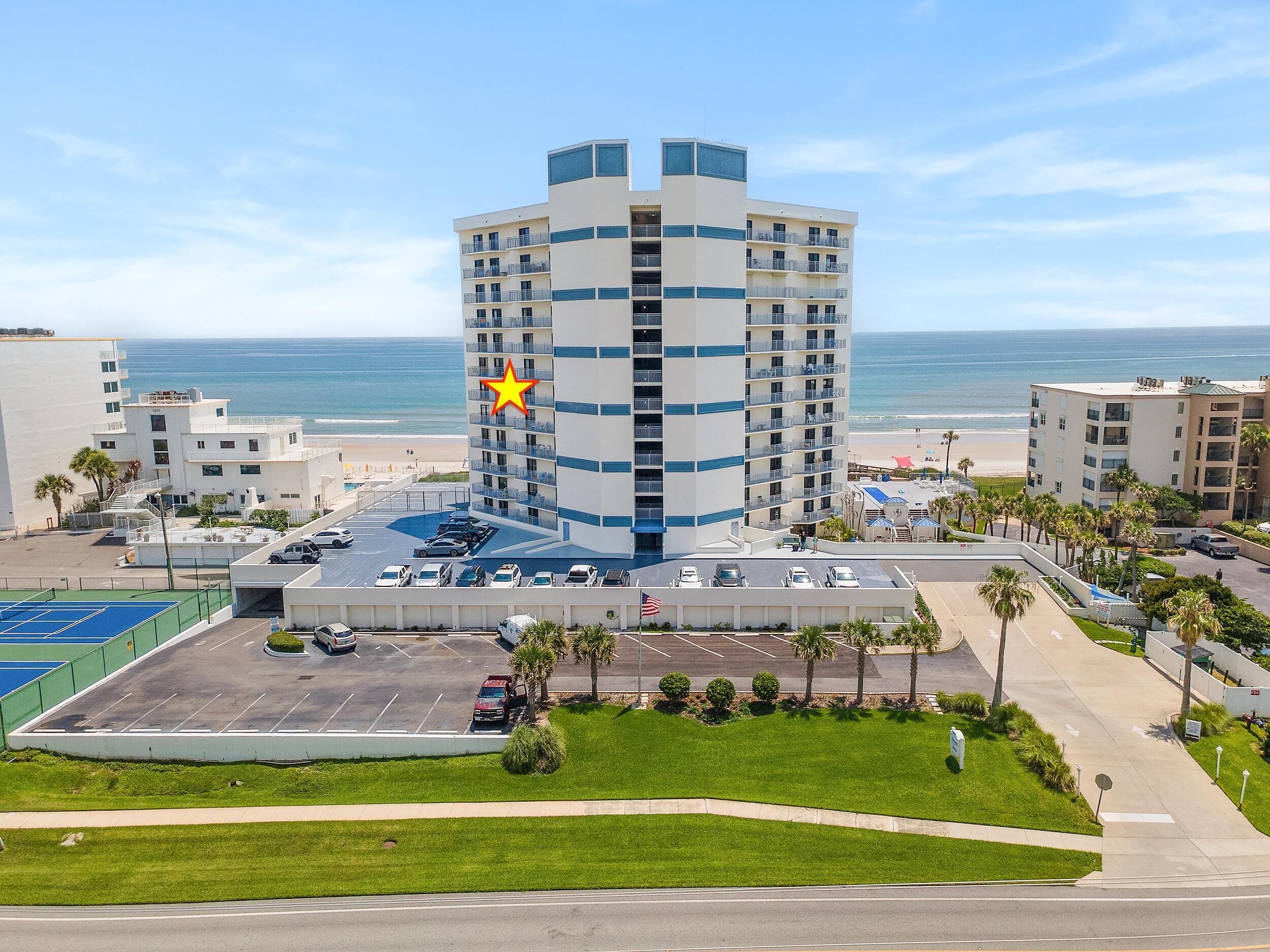 A multi-story hotel with a parking lot, a tennis court, and a view of the beach.