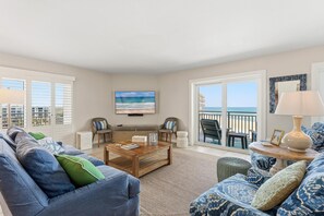 A living room with a large TV, a sofa, a coffee table, and a view of the ocean through a sliding door. - Seascape Towers 621 (New Smyrna Beach)