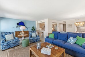Ein Wohnzimmer mit blauem Sofa, gemusterten Sesseln, einem Holztisch und einem Essbereich im Hintergrund.