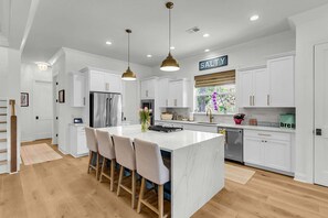 A modern kitchen with white cabinetry, a central island with bar stools, pendant lighting, and a 'SALTY' sign. - 4 Lauden Blvd-Private Saltwater Pool, Grill, 0.4 Miles to Beach (Isle Of Palms)