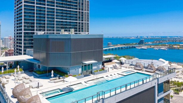 A rooftop pool with lounge chairs and umbrellas, a modern building with a glass facade, and a view of the city skyline and water. - Cielo | Downtown Miami Condo,33 FL,  Rooftop Pool, Gym (Miami)